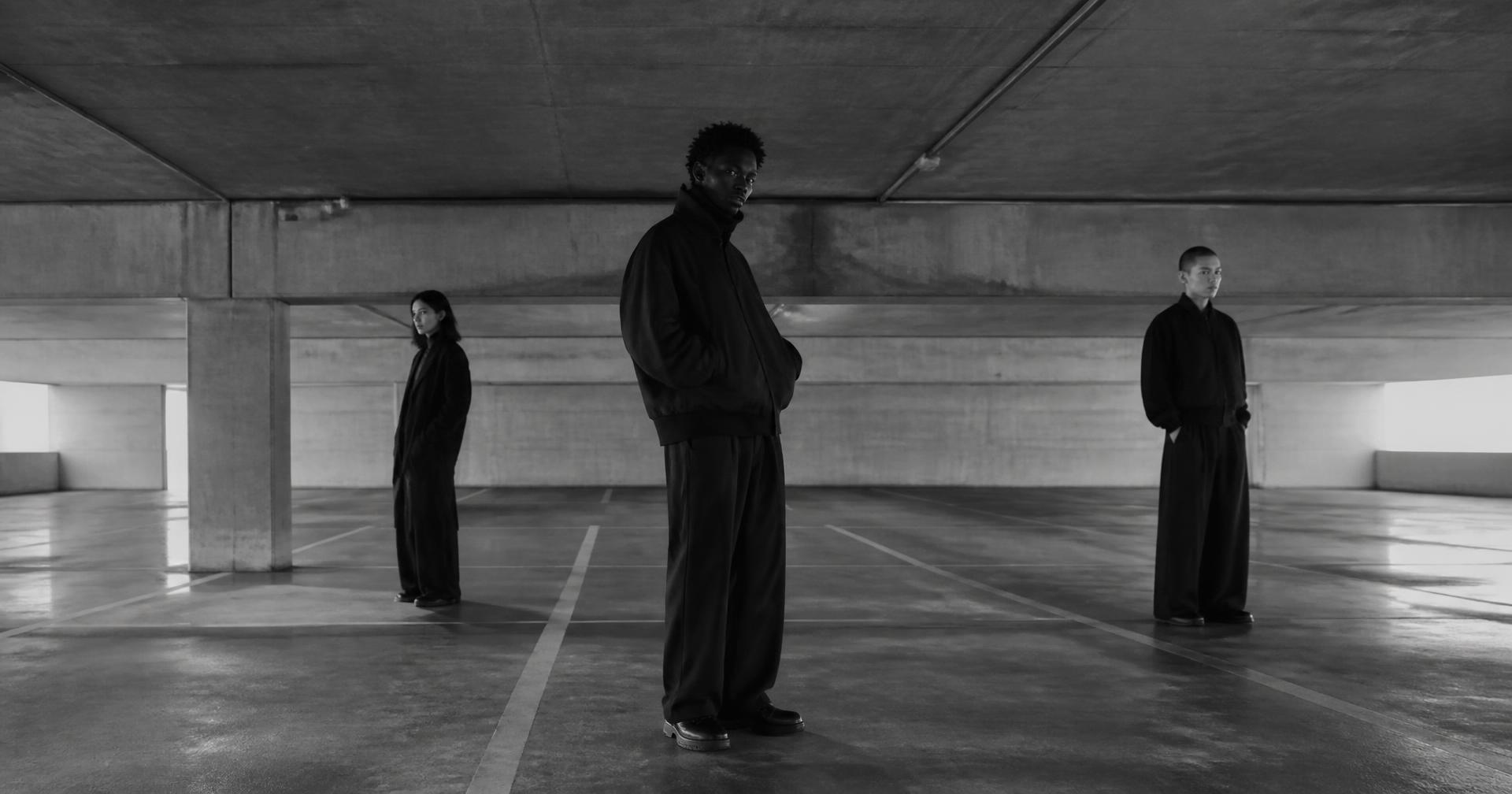 Three figures in monochrome streetwear standing apart in an empty concrete car park